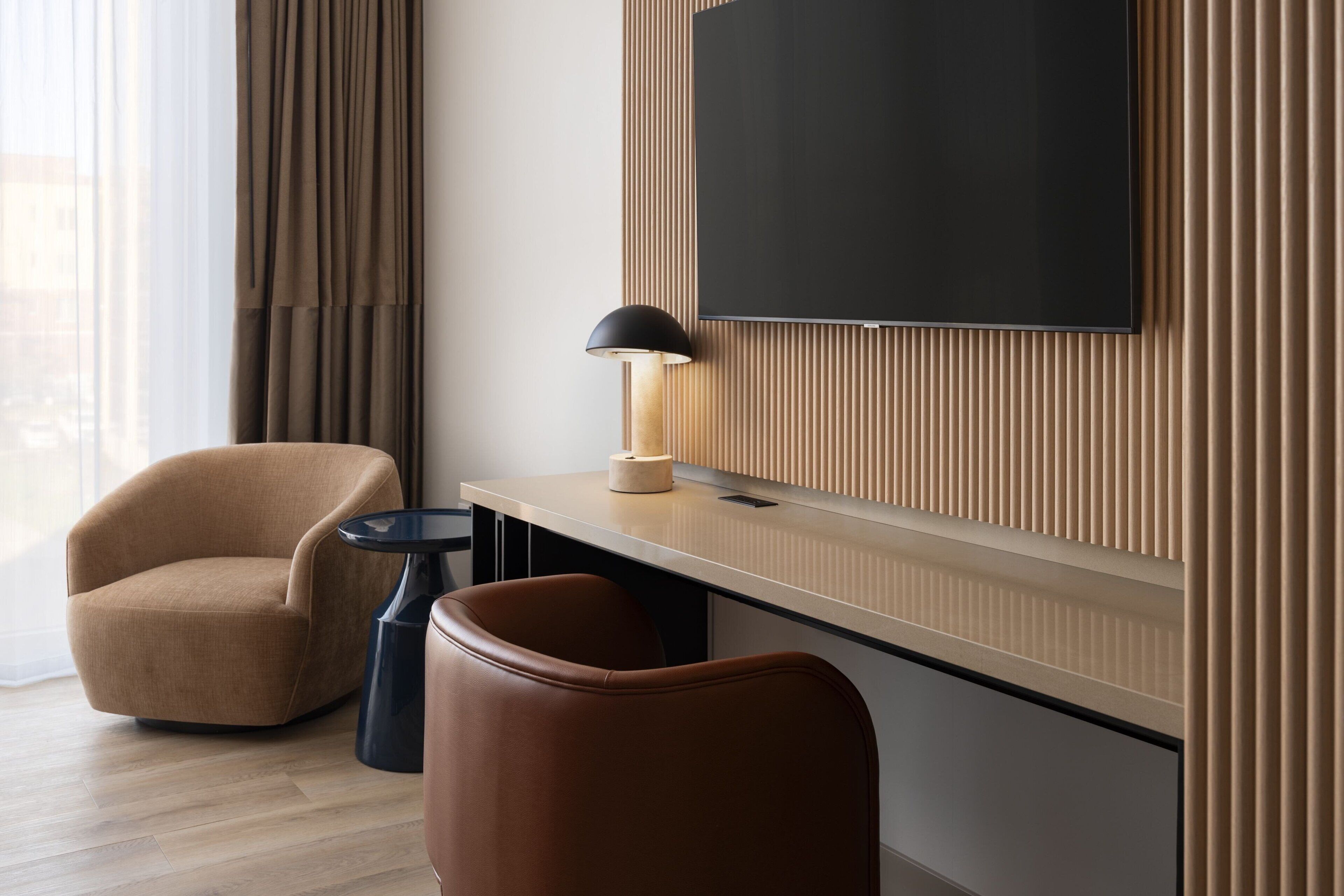 Habitación de hotel moderna con un televisor de pantalla plana grande montado en una pared de paneles de madera, un sillón marrón, una mesita redonda con una lámpara y un reposapiés de cuero.