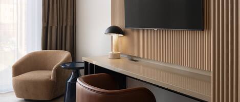 Habitación de hotel moderna con un televisor de pantalla plana grande montado en una pared de paneles de madera, un sillón marrón, una mesita redonda con una lámpara y un reposapiés de cuero.