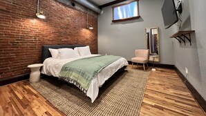 Un dormitorio con pared de ladrillo, cama con manta verde, suelo de madera, ventilador de techo y ventana.