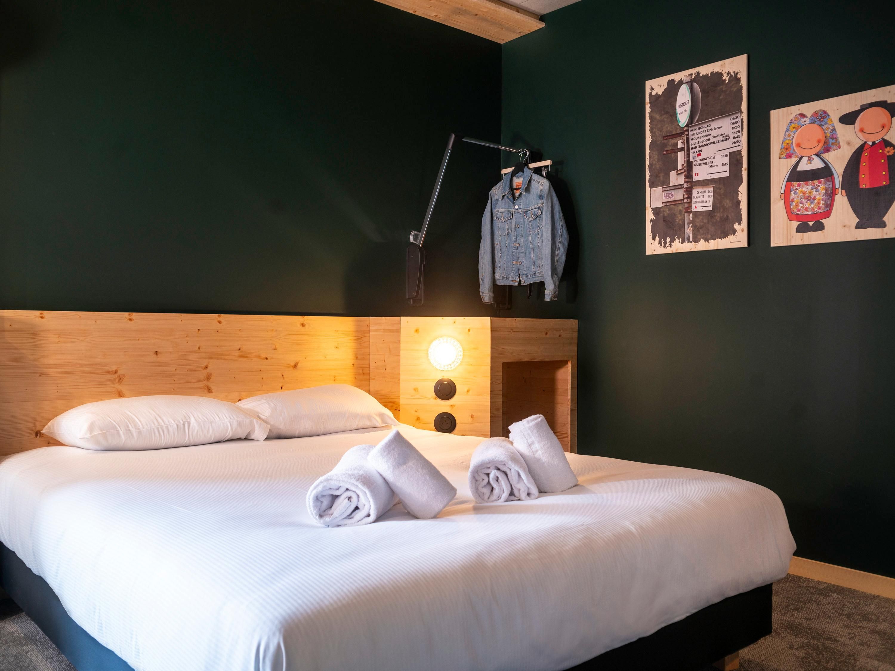A neatly made bed with white linens and rolled towels, a wooden headboard, a wall clock, and a poster of cartoon characters.