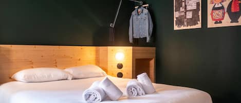 Una cama bien tendida con sábanas blancas y toallas enrolladas, un cabecero de madera, un reloj de pared y un cartel con personajes de dibujos animados.