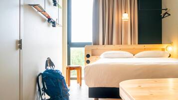 Een slaapkamer met een houten bed, een klein houten krukje, een blauwe rugzak en een raam met gordijnen.