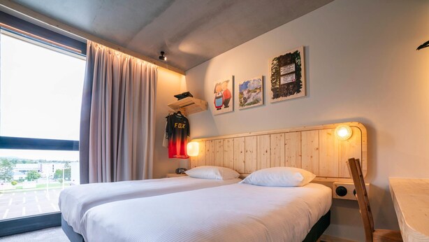 Une chambre d’hôtel avec deux lits, une tête de lit en bois, une fenêtre avec des rideaux et des œuvres d’art accrochées au mur.
