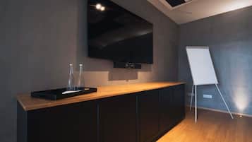 Ein moderner Konferenzraum mit großem, an der Wand montiertem Flachbildfernseher, einem Whiteboard-Ständer und einem Holztisch mit zwei klaren Flaschen.