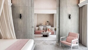 Una habitación de hotel moderna con una cama grande, una zona de estar con sofá y mesa de centro, y vistas a un salón con chimenea.