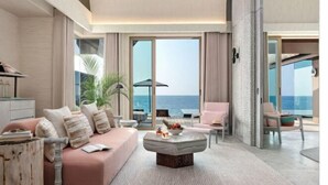 Una sala de estar moderna con sofá, mesa de centro y sillas, ventanas amplias con vista al mar y un balcón con mobiliario exterior.