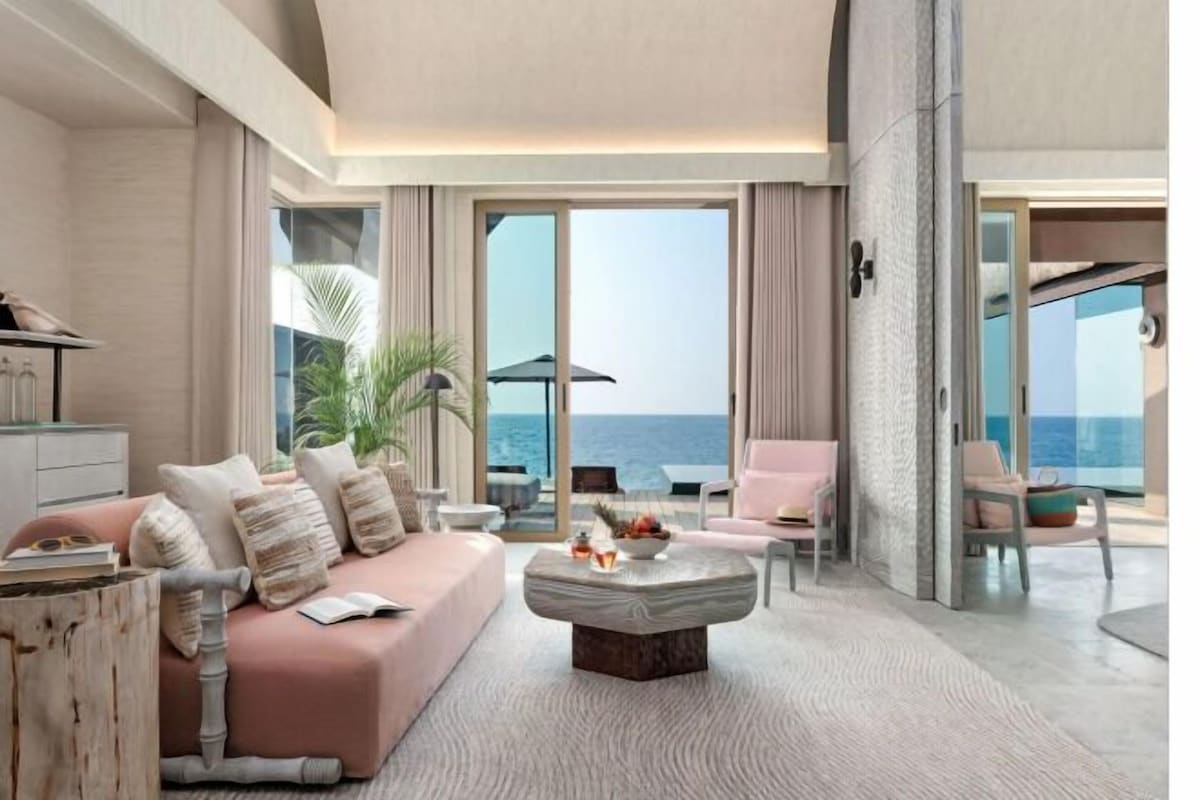 A modern living room with a sofa, coffee table, and chairs, large windows with a view of the sea, and a balcony with outdoor seating.