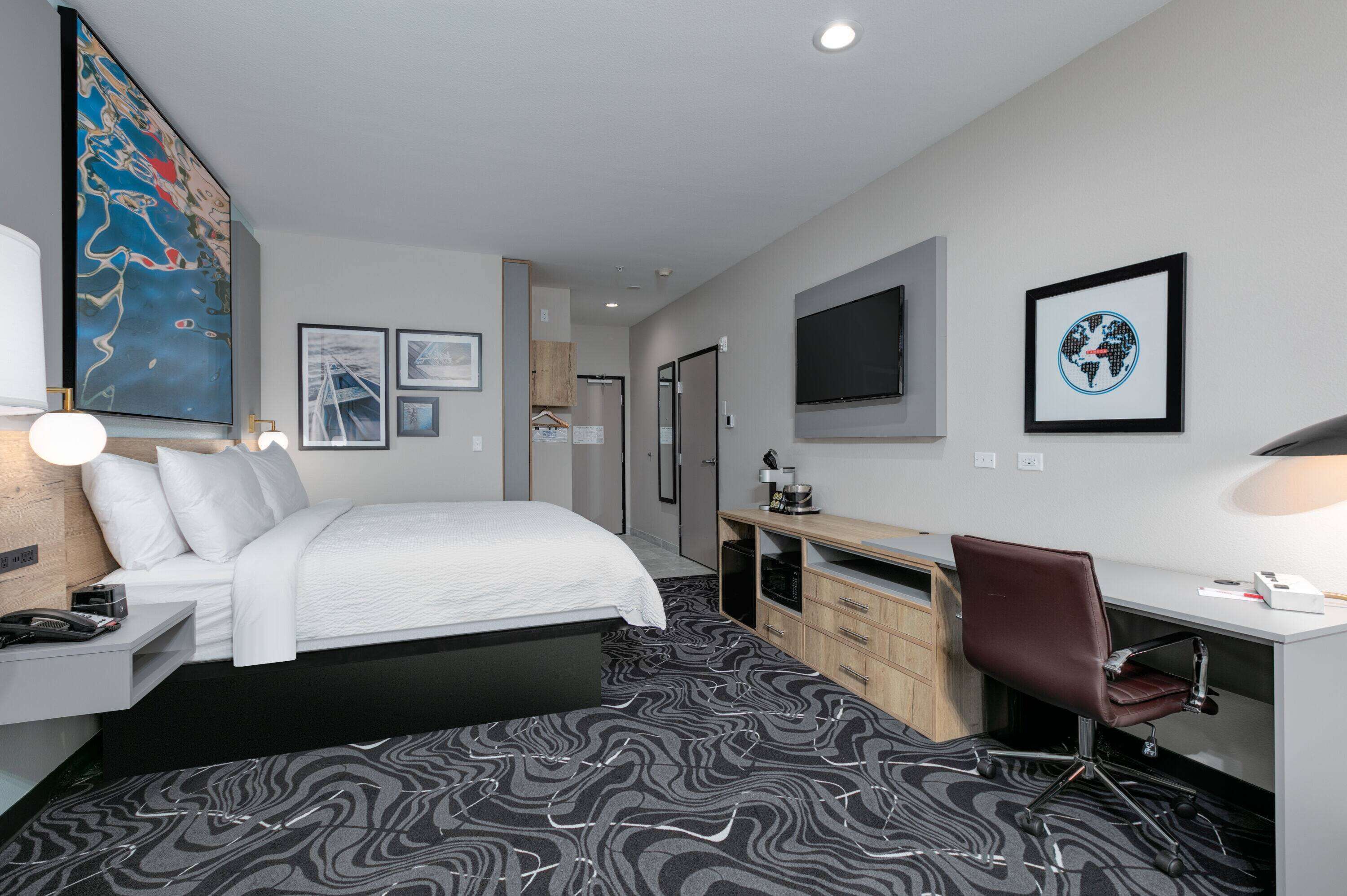 A modern hotel room with a bed, desk, chair, and artwork on the walls.