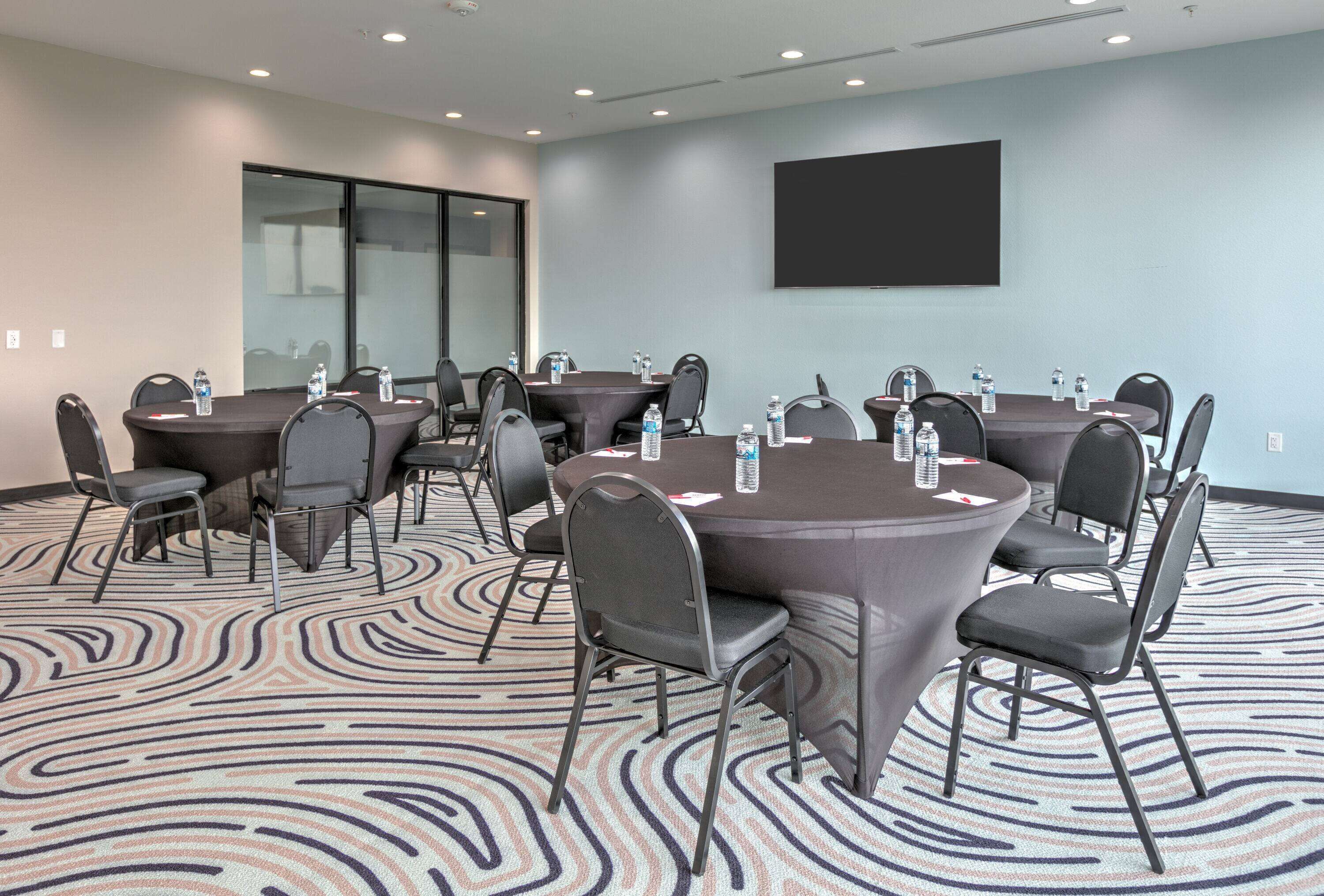 Un salón de conferencias con mesas redondas, sillas, botellas de agua y una pantalla de proyector.