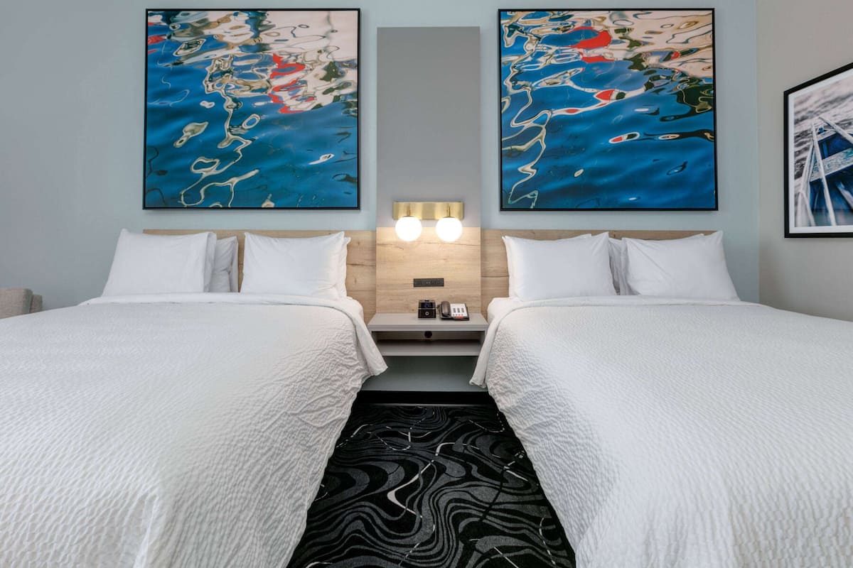 A hotel room with two beds, a nightstand with a phone and a lamp, and abstract paintings on the walls.
