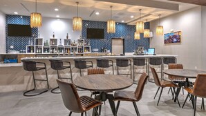 Eine moderne Bar mit verschiedenen Sitzgelegenheiten, darunter Barhocker und runde Tische, und einem an der Wand befestigten Fernsehbildschirm.