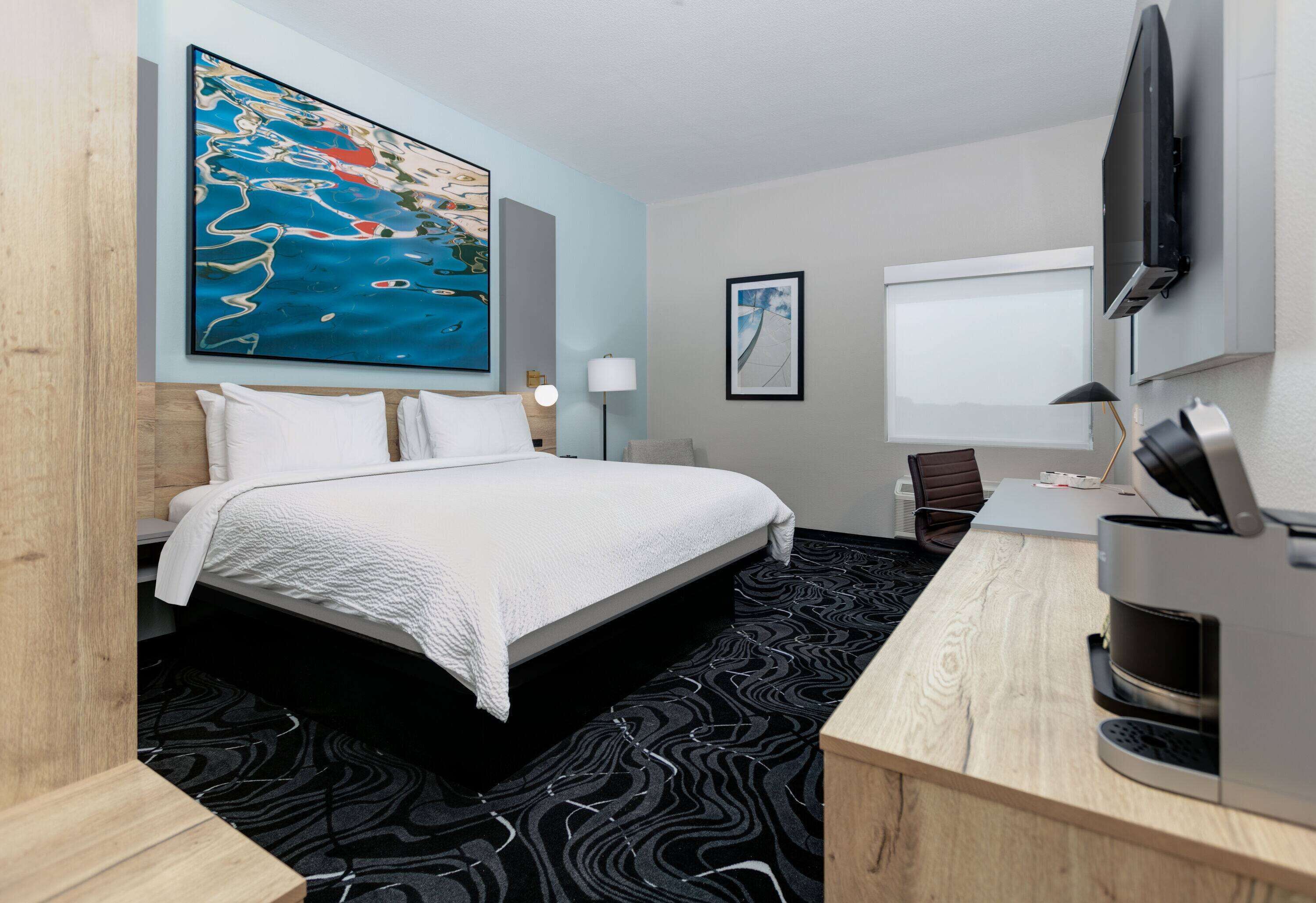 A hotel room with a bed, a desk with a coffee machine, a TV, and a large abstract painting on the wall.