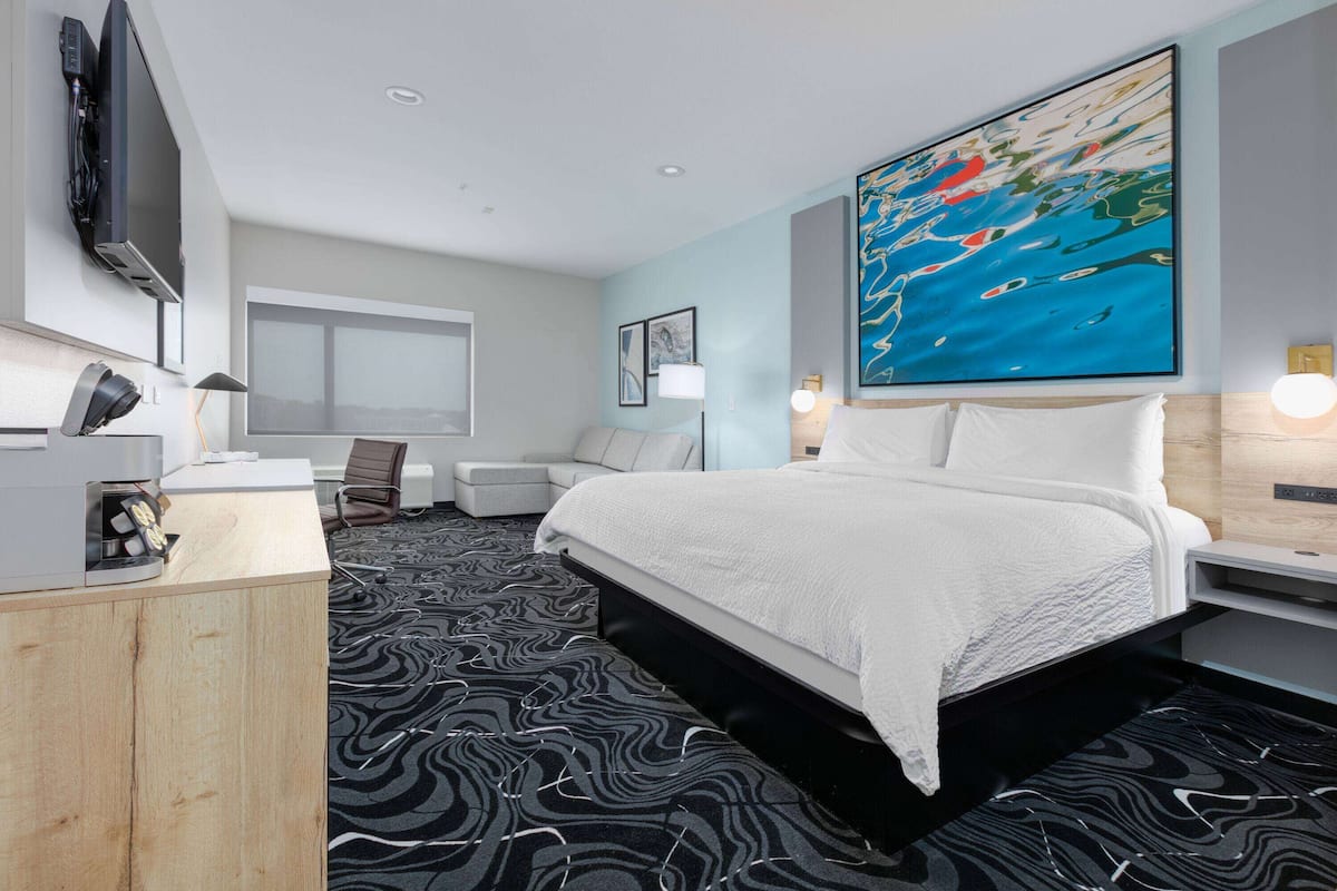 A modern hotel room with a large bed, a desk, a chair, a flat-screen TV mounted on the wall, and a large abstract painting above the bed.