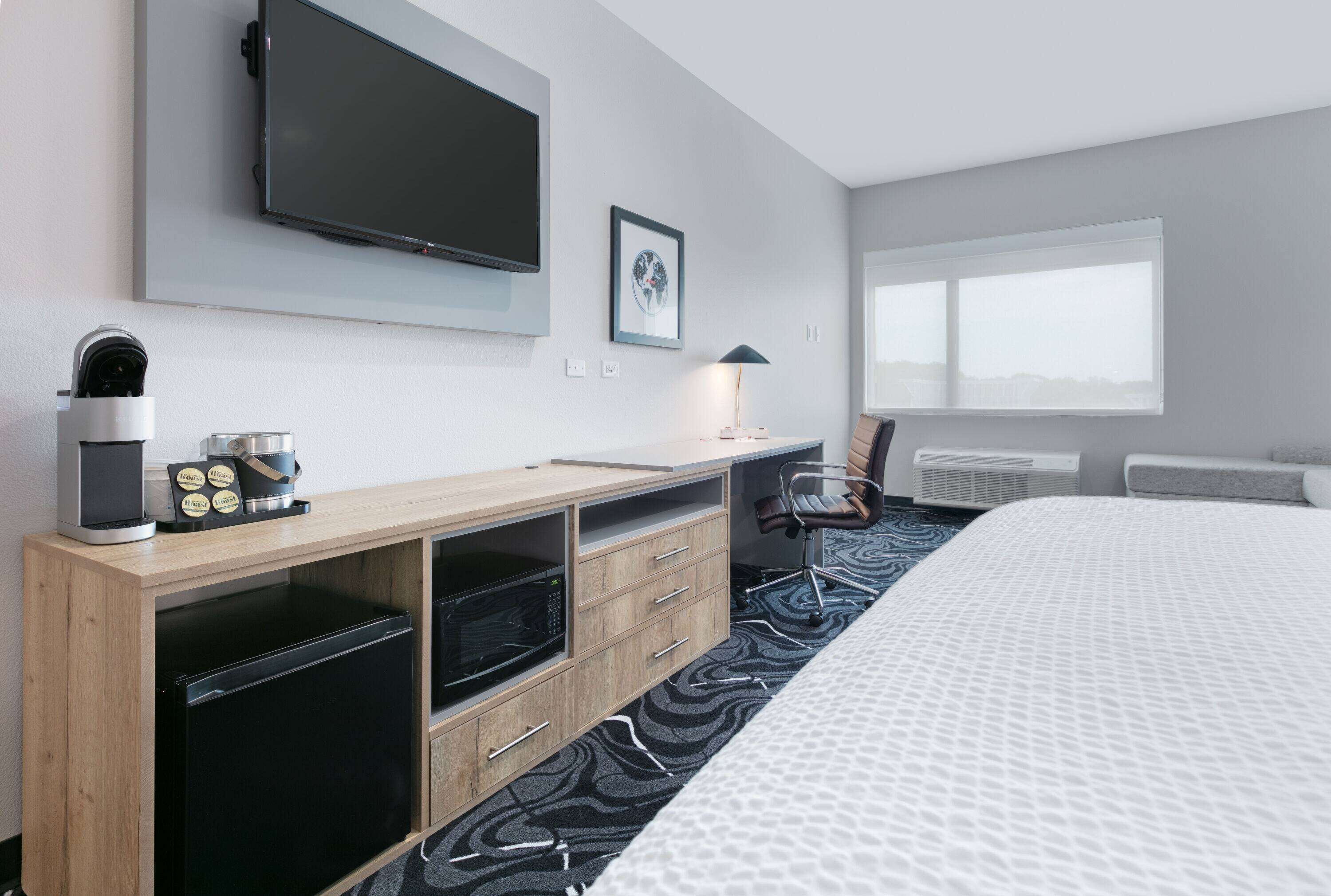 A hotel room with a bed, a TV mounted on the wall, a desk with a microwave, a chair, and a window with a view.