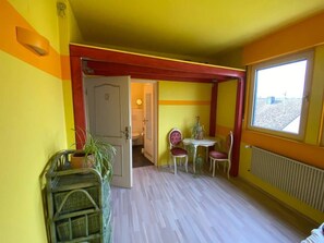 Quarto com decoração em amarelo e vermelho, uma pequena mesa com cadeiras, uma planta em vaso e uma janela com vista.