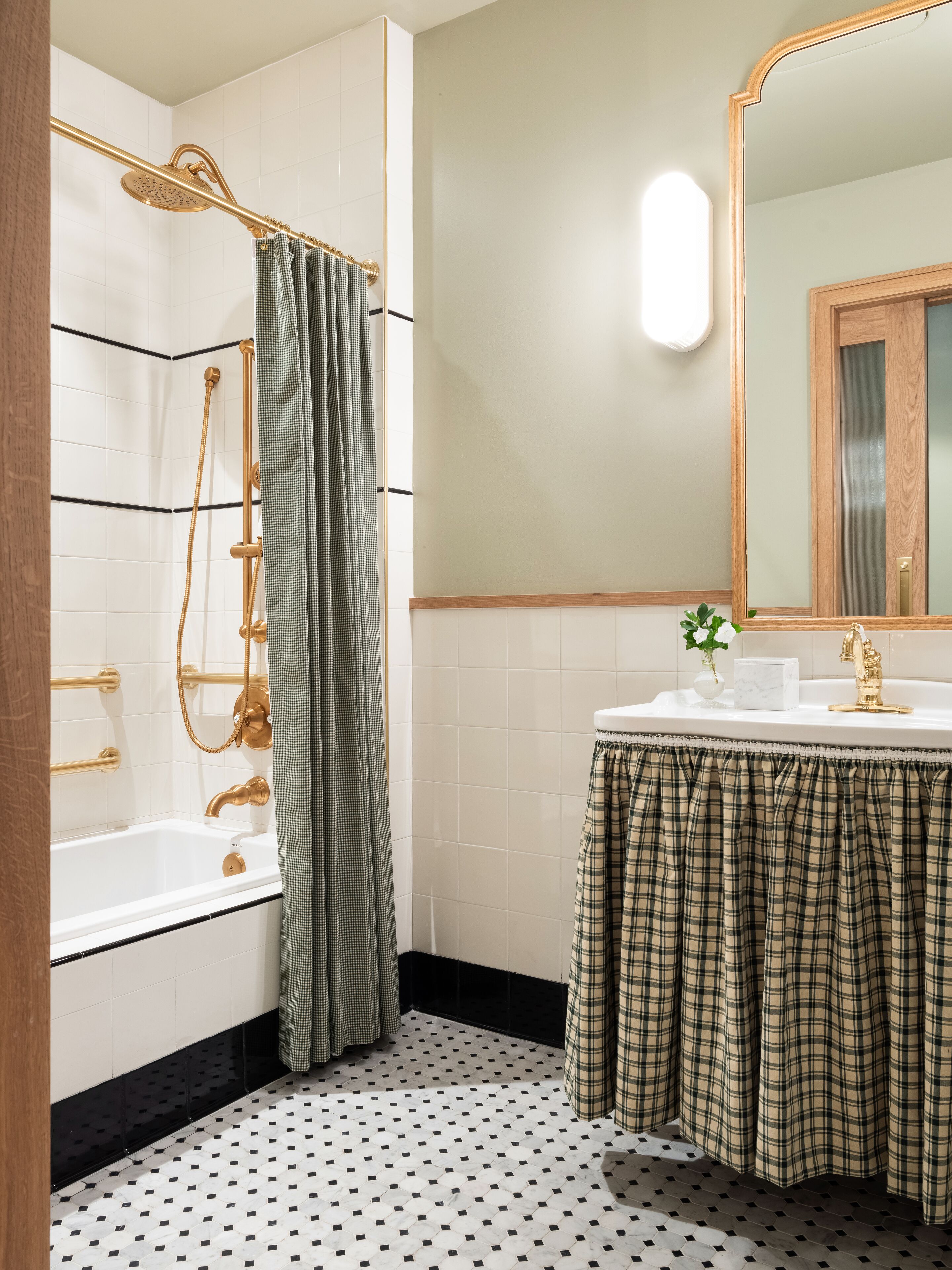 Un baño con una bañera blanca, un cabezal de ducha dorado, una cortina de ducha verde a cuadros, un lavabo blanco con grifo dorado y un espejo con marco de madera.