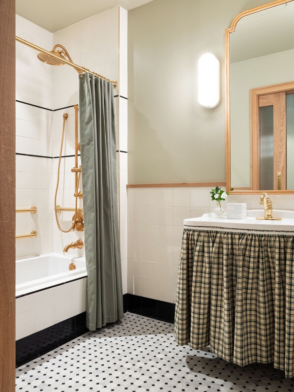 Un baño con una bañera blanca, un cabezal de ducha dorado, una cortina de ducha verde a cuadros, un lavabo blanco con grifo dorado y un espejo con marco de madera.
