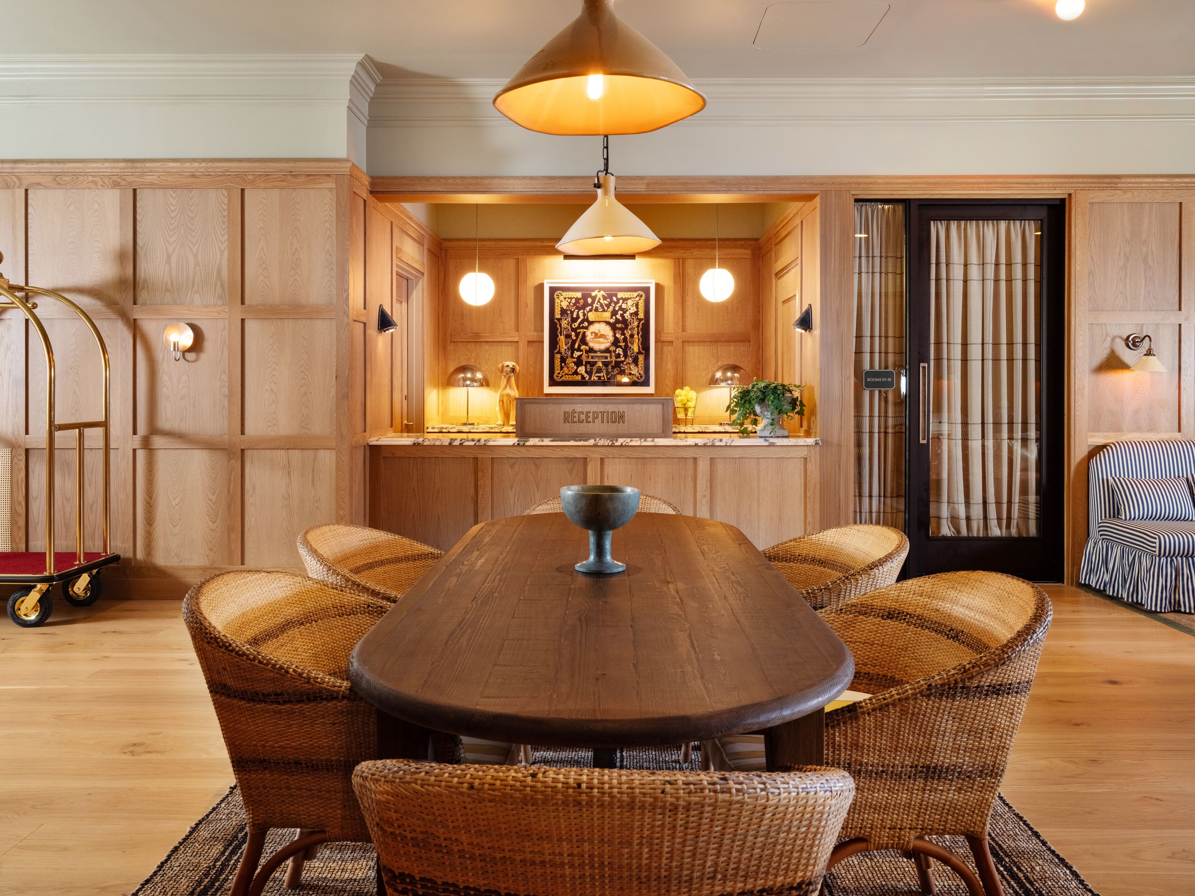 A wooden-paneled room with a central dining table, wicker chairs, a reception desk, and a striped armchair.