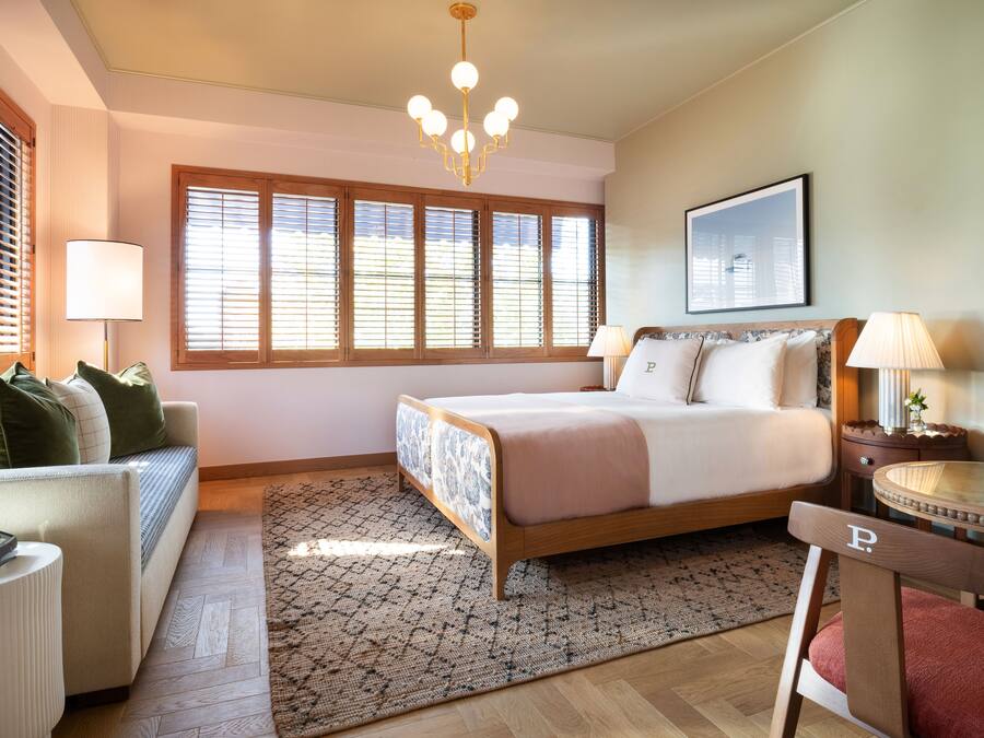 Ein Schlafzimmer mit einem Bett, einem Sofa, einem Sessel, einem Schreibtisch und einem Fenster mit Jalousien.