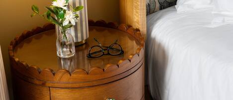 A wooden bedside table with a lamp, a vase with flowers, and a pair of glasses. There is a bed with white bedding and a patterned headboard.