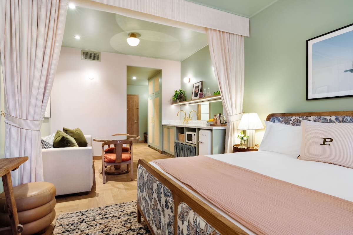 A hotel room with a bed, a desk, a chair, a small table, and a kitchenette.