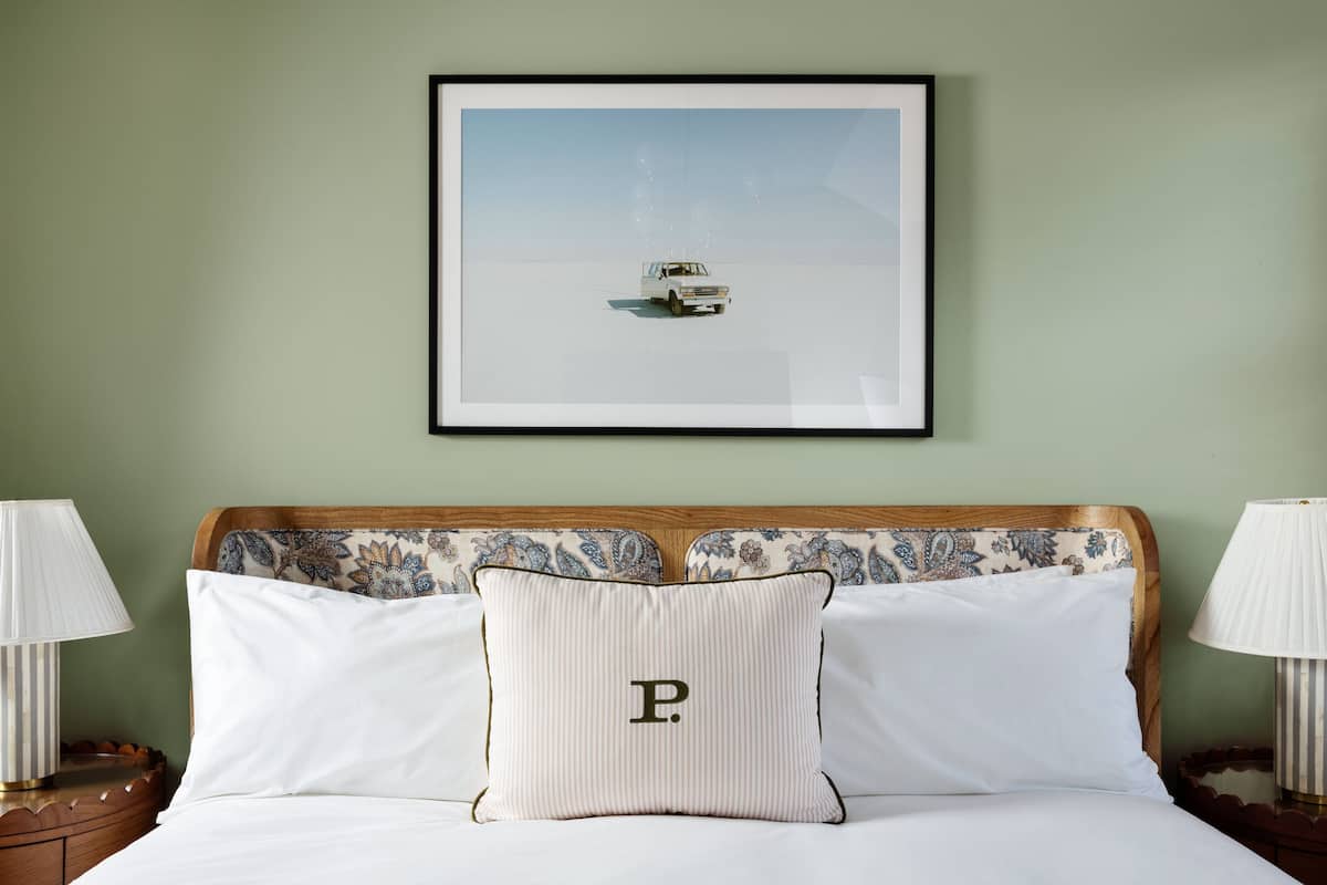 A neatly made bed with a pillow featuring a large letter 'P', a framed picture of a boat on water, and two bedside lamps.