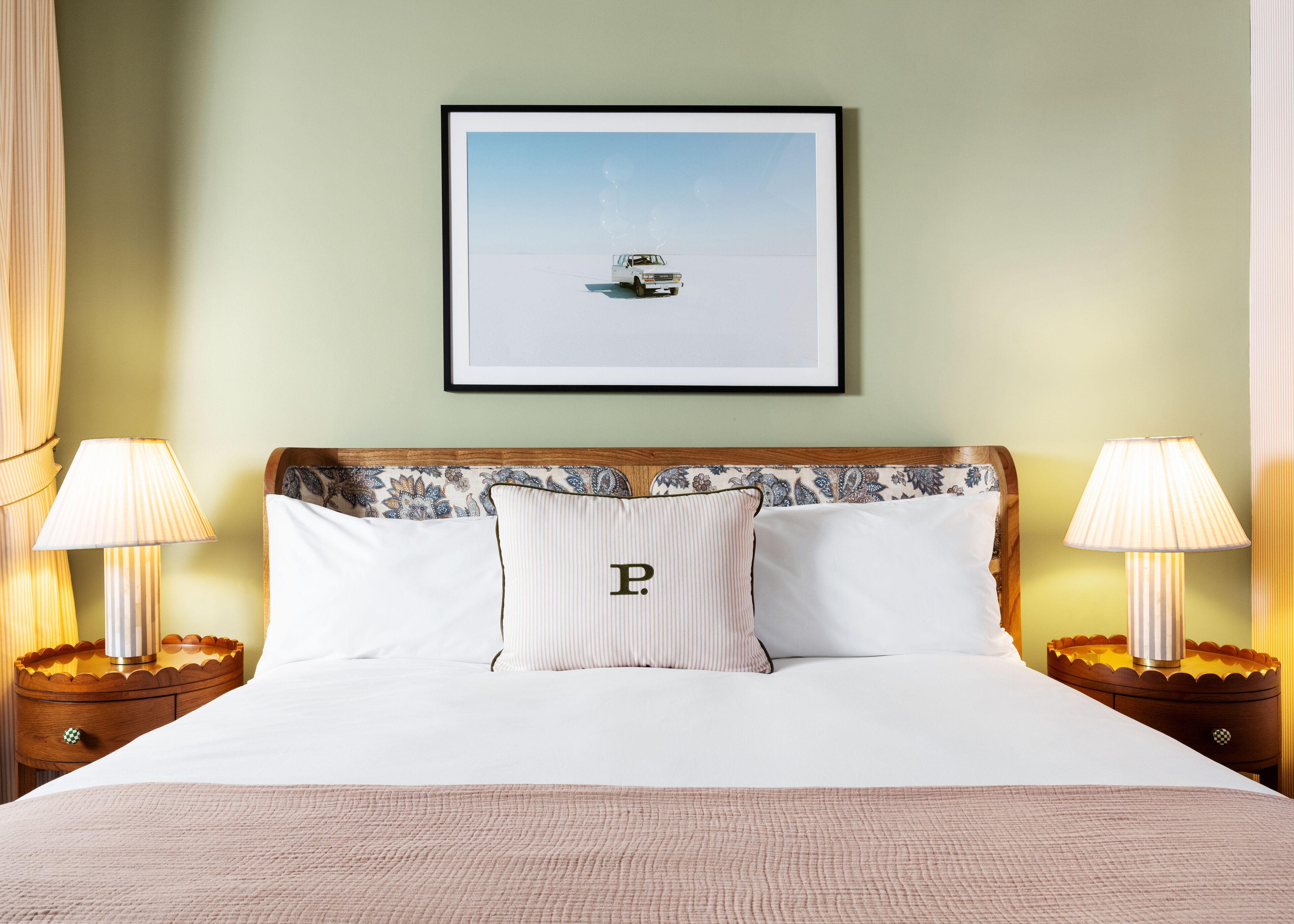 A neatly made bed with a pillow featuring a capital 'P', two bedside lamps, and a framed picture of a car on the wall.