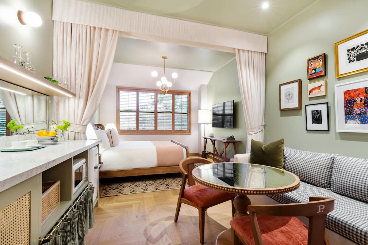 A compact hotel room with a bed, a dining area with a table and chairs, a sofa, a TV, and artwork on the walls.