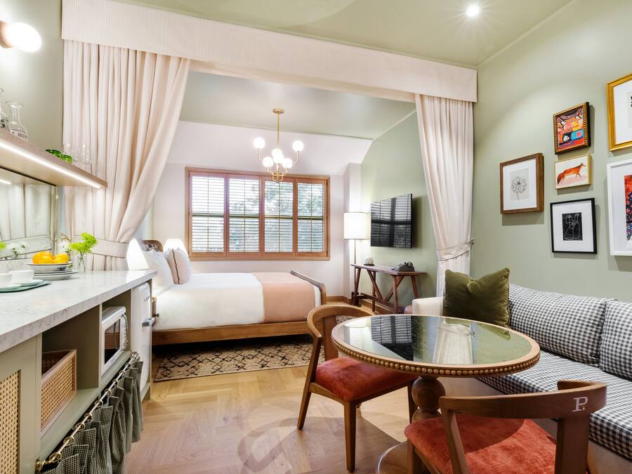 A compact hotel room with a bed, a dining area with a table and chairs, a sofa, a TV, and artwork on the walls.