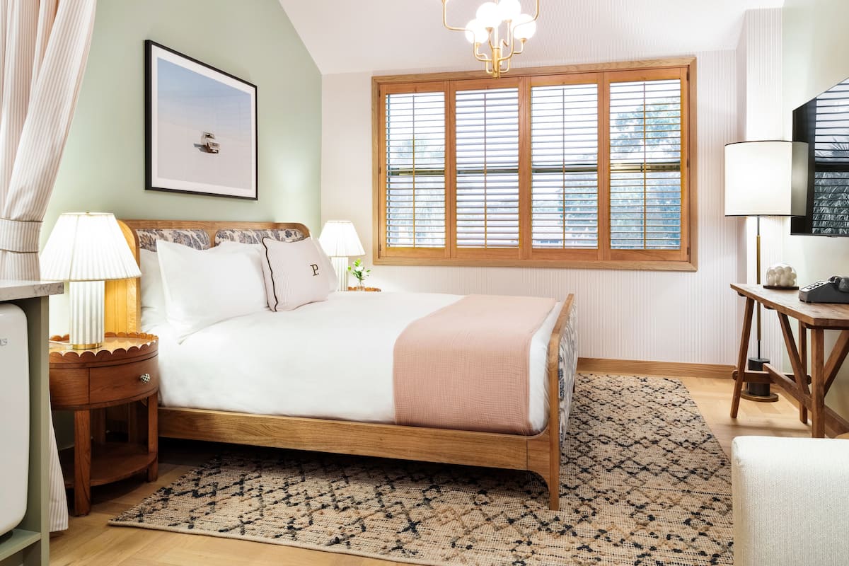 A bedroom with a bed, bedside tables, a chandelier, a window with blinds, a television, and a framed picture on the wall.