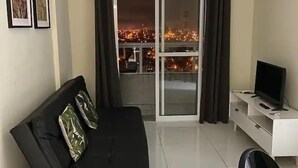 A modern living room with a black sofa, a TV on a white stand, and a view of a cityscape through a sliding glass door. - Apto com vista pro Mar sacada gourmet (Praia Grande)