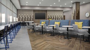 A modern restaurant with long communal tables, individual seating, and a large screen wall. - Home2 Suites By Hilton Cullman (Cullman)