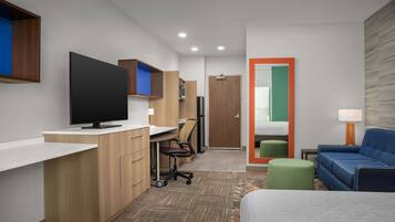 A modern hotel room with a flat-screen TV, wooden cabinets, a desk, a chair, a sofa, a green ottoman, and a mirror with a colorful frame.