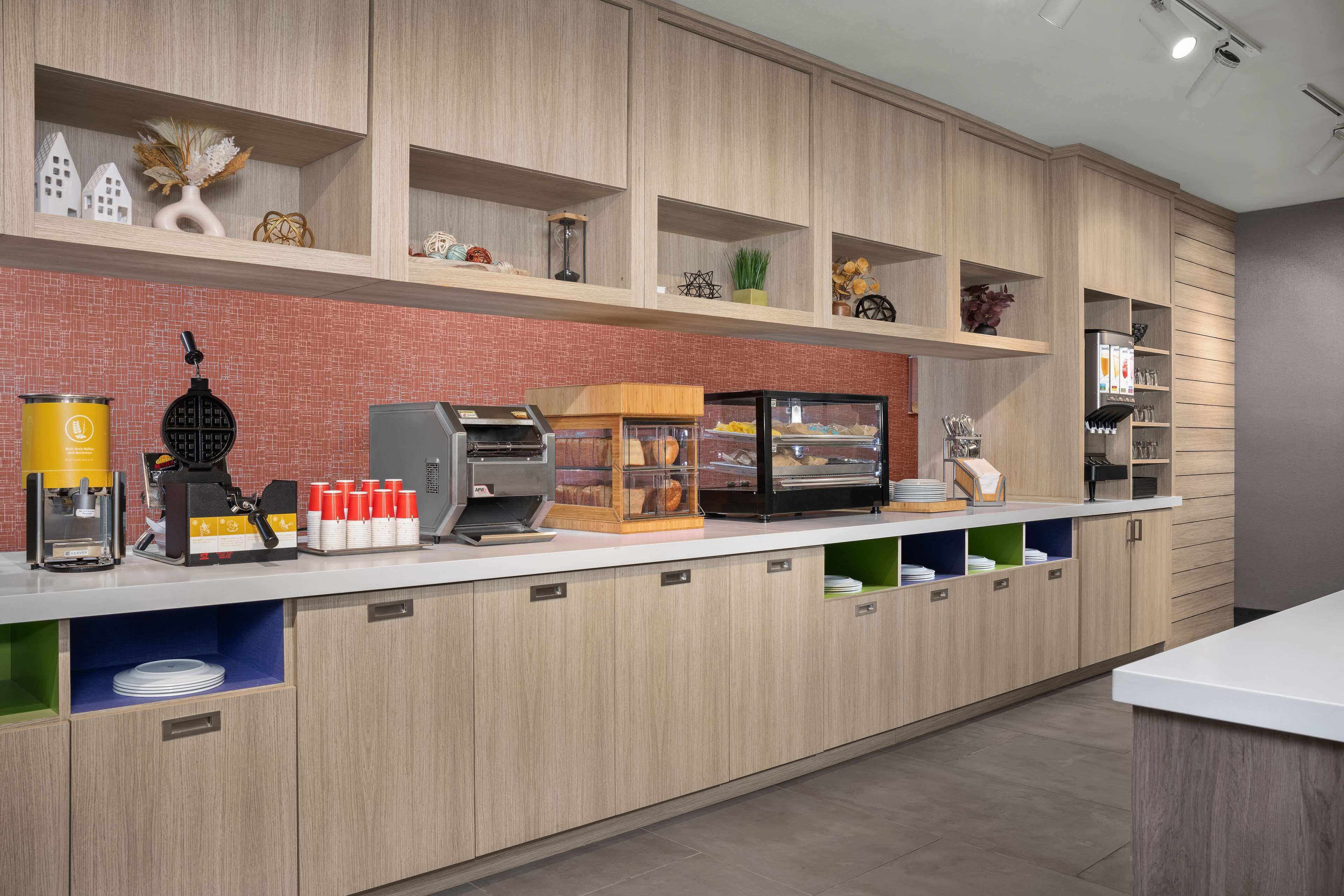 Un área de cocina moderna con gabinetes de madera, protector contra salpicaduras de ladrillo y varios electrodomésticos, incluyendo una cafetera y un expositor para pan.
