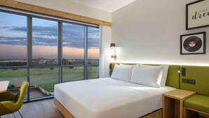 Habitació d'hotel moderna amb una finestra gran, un llit, un escriptori i un sofà verd.