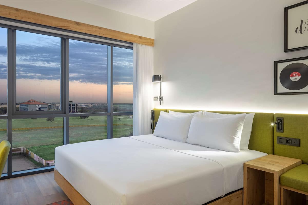 A modern hotel room with a large window, a bed, a desk, and a green sofa.