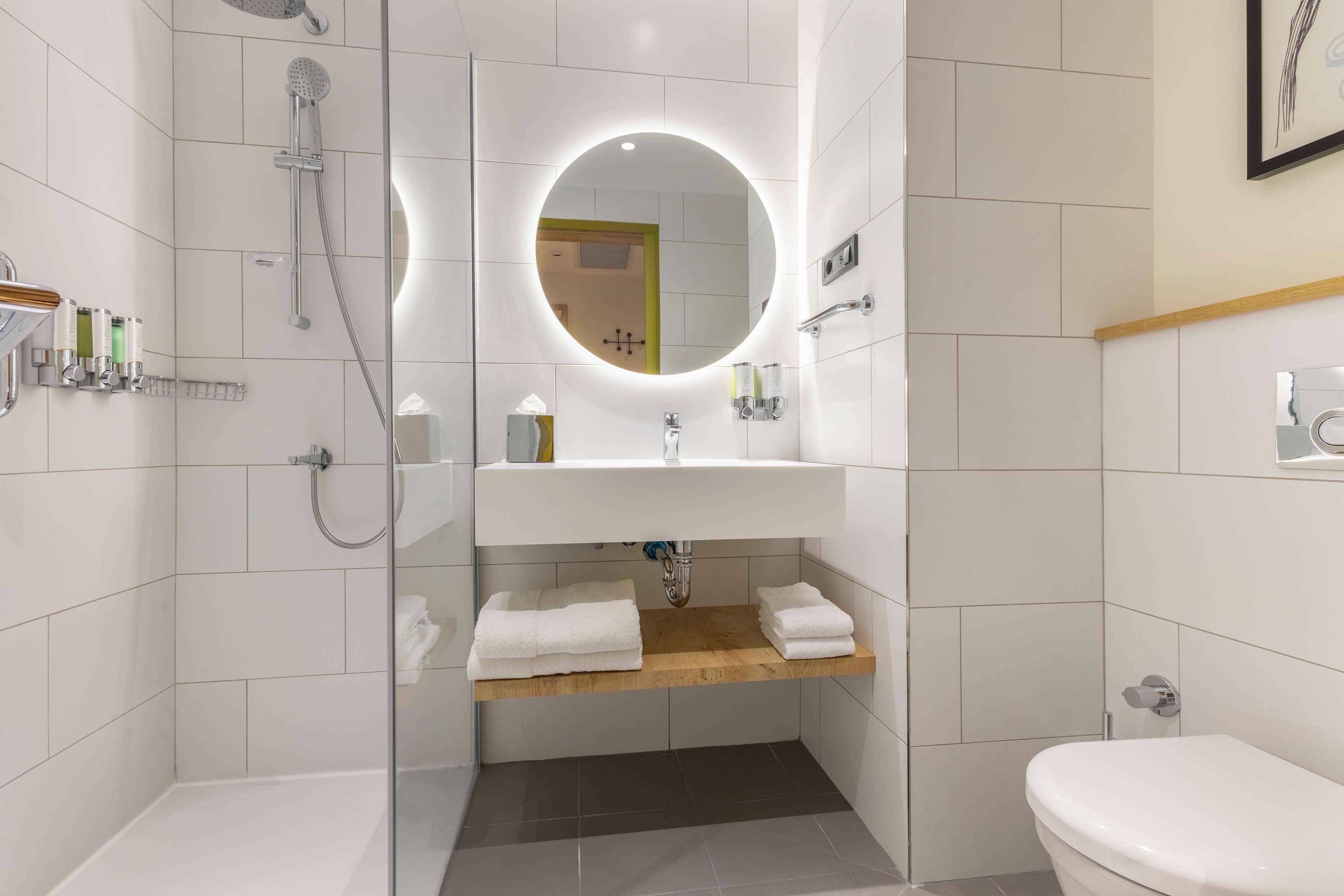 Un baño moderno con ducha envuelta en vidrio, un espejo redondo, un estante de madera con toallas y un lavabo blanco.