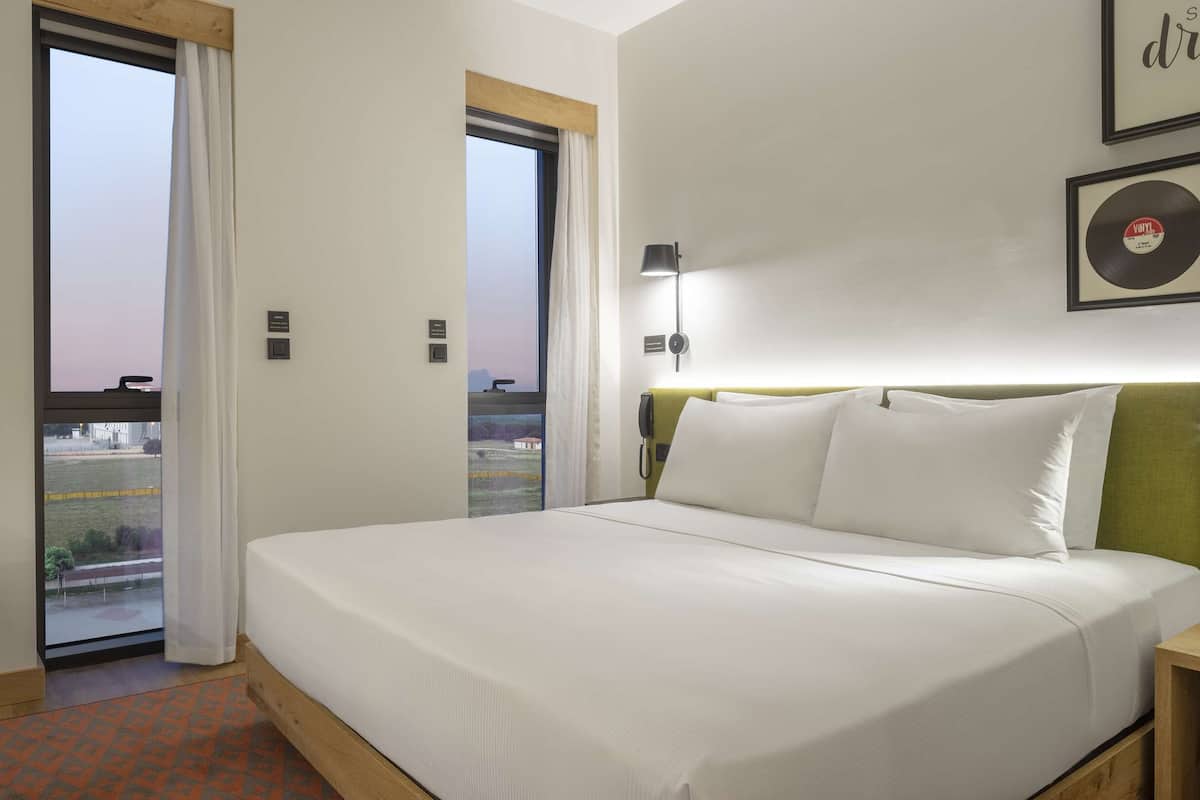 A hotel room with a bed, a desk, a chair, a bedside table, a lamp, and a view of the outside.
