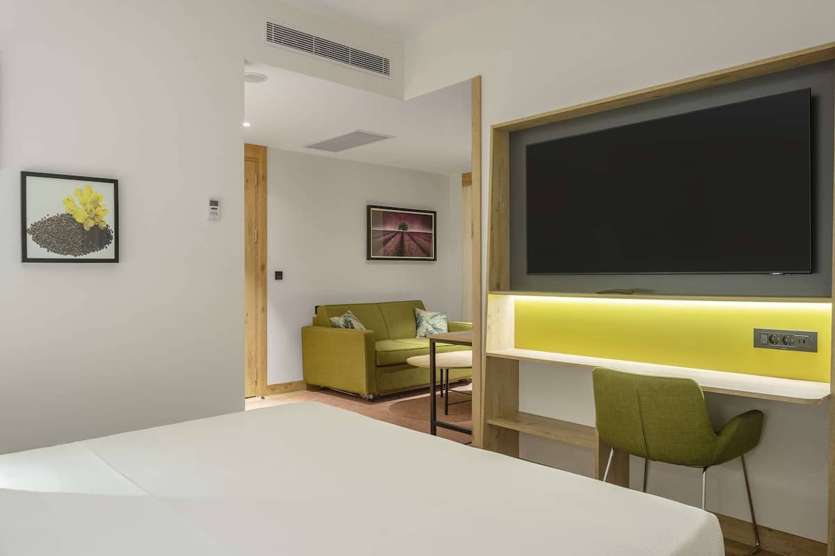 A hotel room with a bed, a desk with a chair, a TV, and framed pictures on the wall.