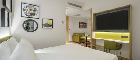 A hotel room with a bed, a desk with a chair, a TV, and framed pictures on the wall.