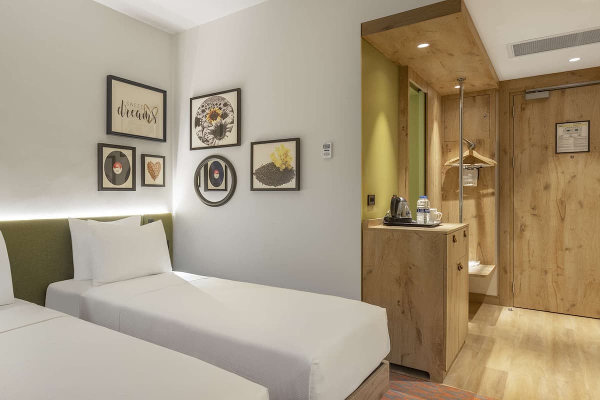 A hotel room with two beds, a bedside table with a lamp, a wooden wardrobe, and wall-mounted artwork.