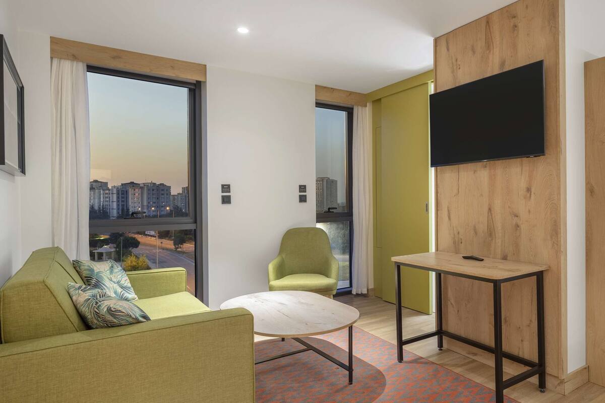 A modern hotel room with a sofa, a small round table, a desk with a chair, a TV mounted on a wooden panel, and large windows offering a city view.