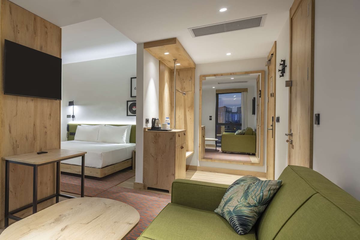 A compact hotel room with a bed, a small desk, a sofa, and a mirror reflecting a cityscape.