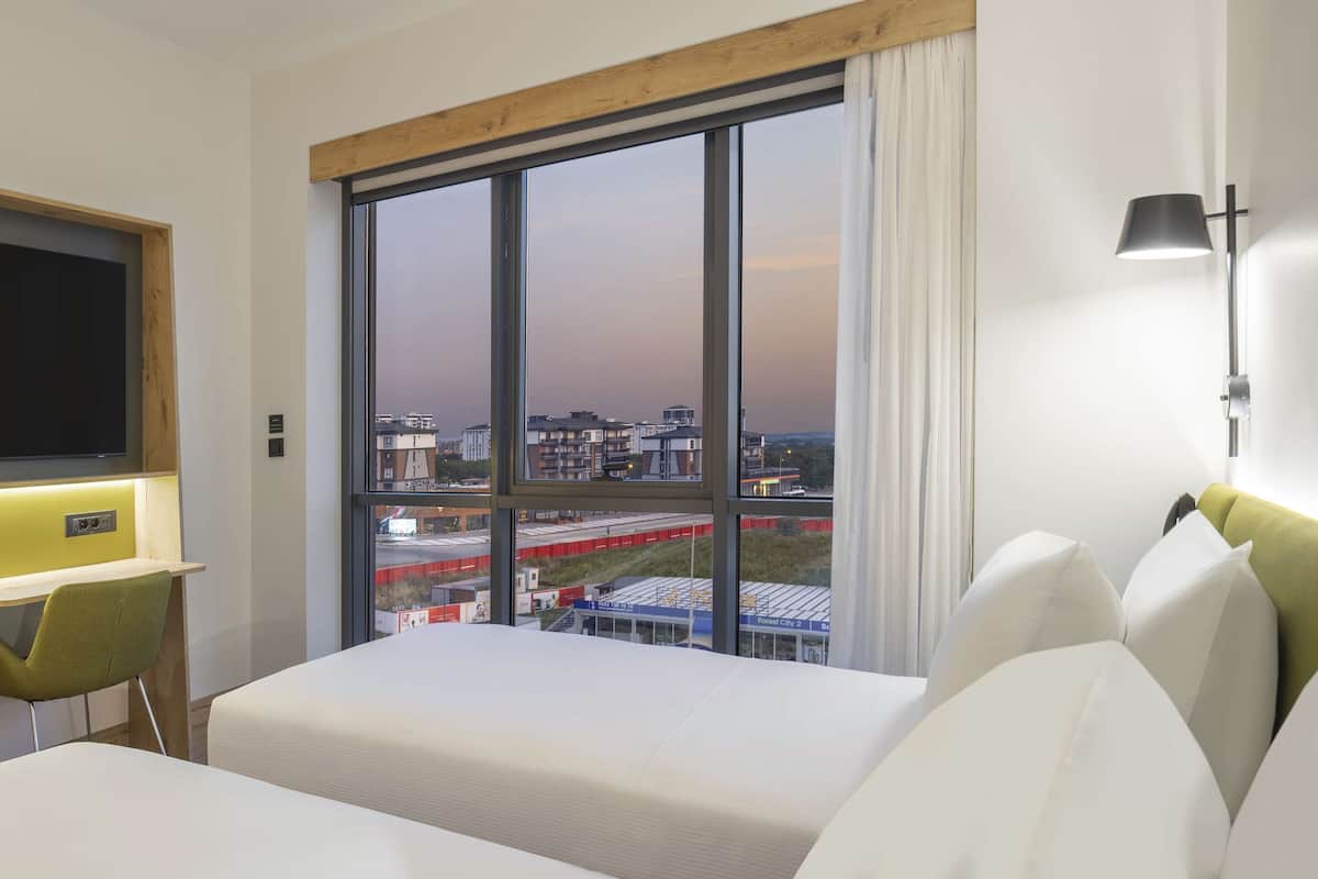A hotel room with a large window, a flat-screen TV, a desk, and two beds.
