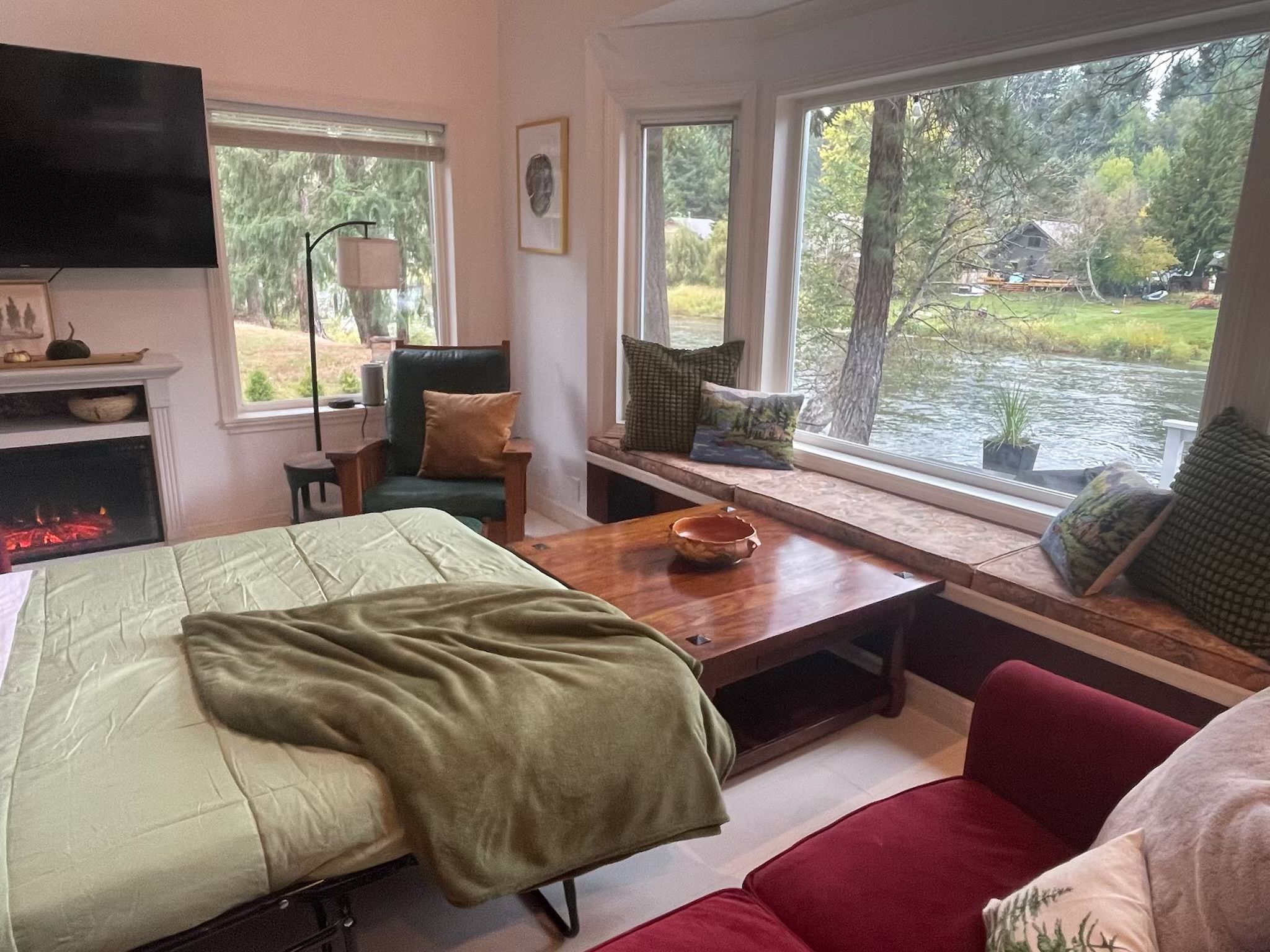 A cozy room with a bed, a desk, a chair, and a couch. There are windows with a view of a river and trees.