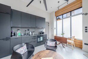 A modern kitchen with dark cabinets, a built-in oven, and a dining area with a wooden table and chairs. - Stylishly Designed 1BDR Flat by Battersea Park (London)