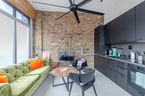 A modern kitchen with a brick wall, a ceiling fan, and a dining area with a green sofa and a coffee table. - Stylishly Designed 1BDR Flat by Battersea Park (London)
