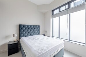 A bedroom with a large window, a bed with a white duvet, a dark blue headboard, and a small bedside table with a lamp. - Stylishly Designed 1BDR Flat by Battersea Park (London)
