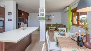 Eine moderne Küche mit weißer Arbeitsplatte, Holzschränken und einem eingebauten Ofen. Ein Essbereich mit Tisch für zwei Personen, einem grünen Sofa und einem Fenster mit Blick auf Grünflächen.