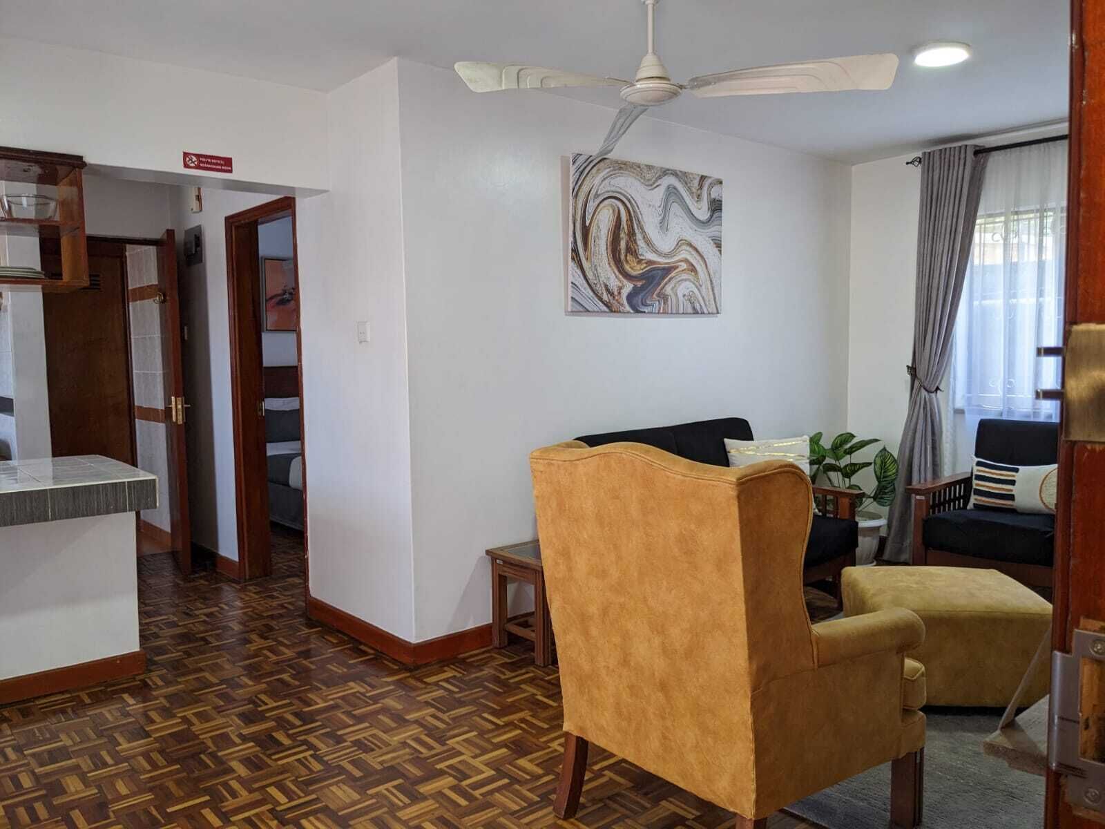 Una habitación con piso de madera tipo espiga, ventilador de techo, una obra de arte en mármol, sofá negro, sillón amarillo y una ventana con cortinas.